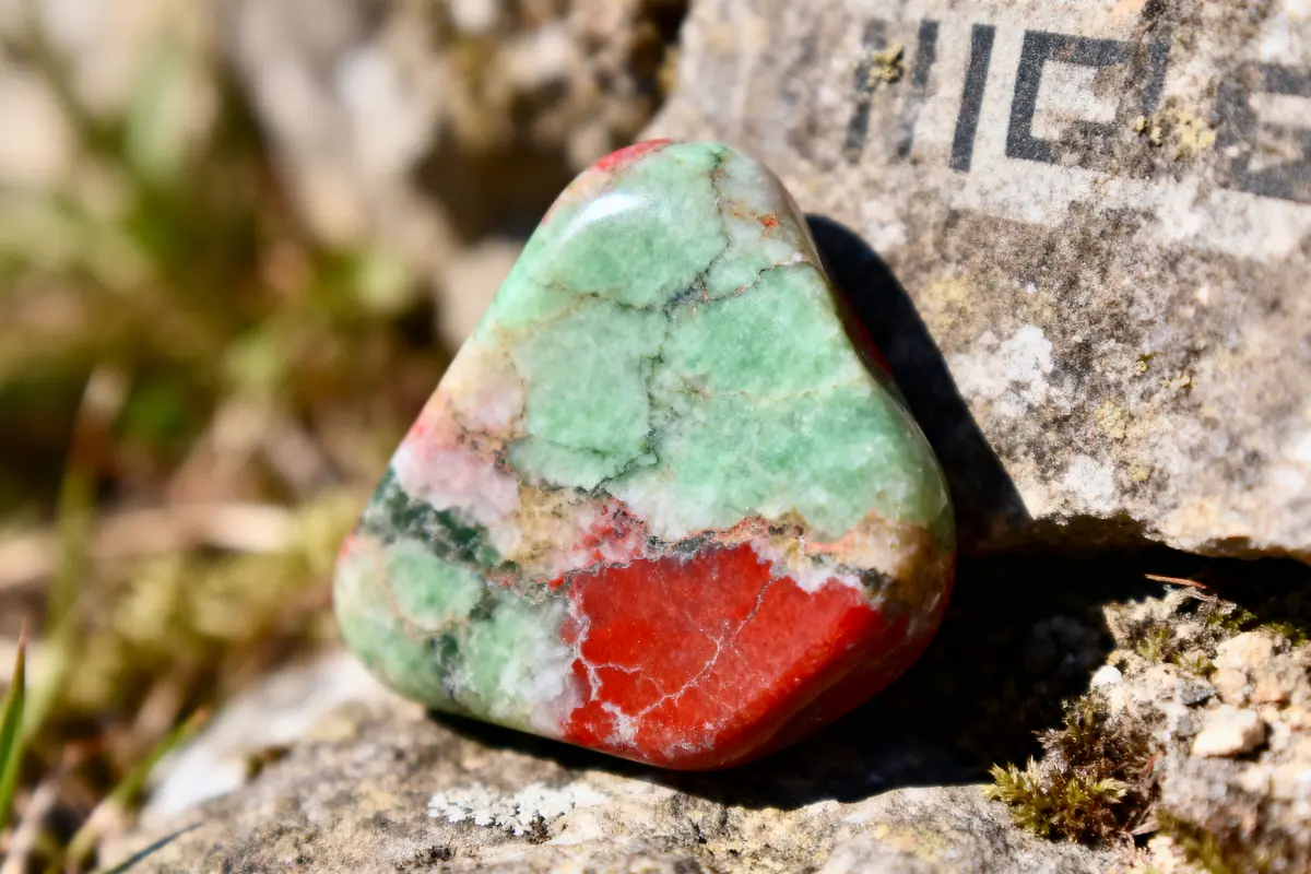Pierre polie aux couleurs verte et rouge, posée sur un rocher gris avec inscriptions partiellement visibles.