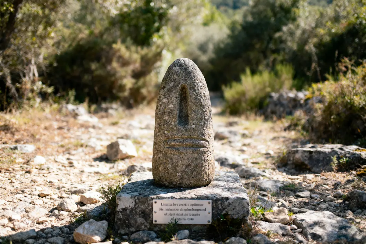 Petite sculpture en pierre sur socle, entourée de végétation et rochers, plaque métallique au bas.